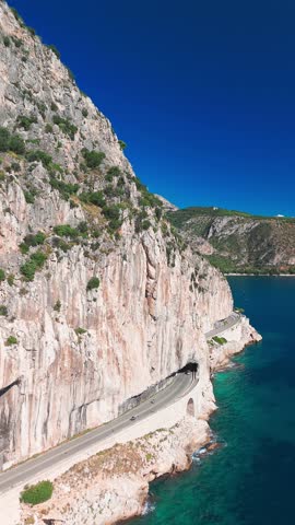 Aerial view of the French Riviera coastline between Nice and Monaco. A car drives along the Corniche Inferieure road, winding through high cliffs above the turquoise waters of the Mediterranean Sea