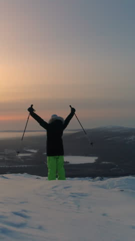 AERIAL: Flying around woman in warm winter ware raising arms on top of snowy mountain. Female traveler standing on top of the mountain on snowshoes, raising arms victoriously. Active people in winter