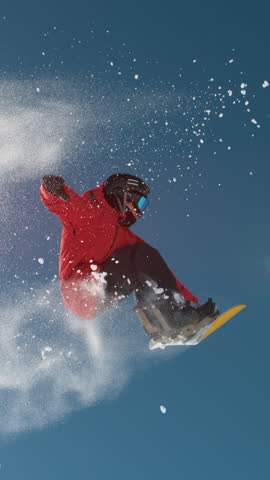 SLOW MOTION CLOSE UP: Snowboarder jumping big air kicker, spraying snowflakes and flying over sun on perfect winter day. Snowboard jump in snow park. Sunbeams shining past jumping boarder in mountains