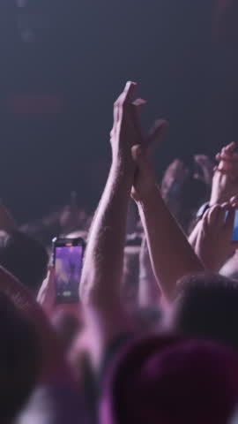 Vertical video: Waving fans hands at band concert in magenta stage light spotlights. Close up, happy people clap their hands, film show on phones. Disco party illumination by pink light. Crowd dance