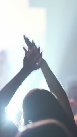 Vertical video: Back view, happy woman fan dancing at band concert, raising hands up in bright spotlight. Girl silhouette waving hands in people crowd on stage club background. Music festival party