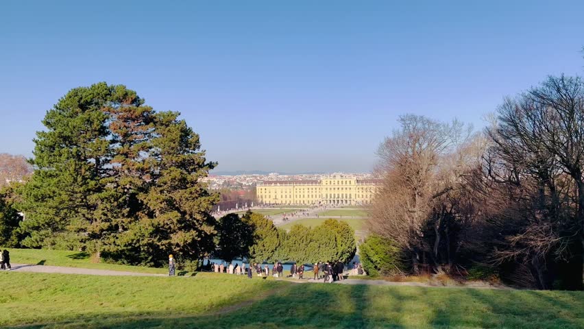 Schönbrunn imperial palace from Gloriette hill in Vienna, Austria during sunny fall, winter day. Locals and tourists walking and jogging around gravel path, nature park full of trees and green grass.