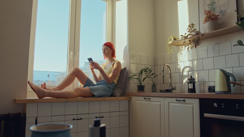 Young woman with bright red hair sitting on sunlit windowsill in cozy kitchen, looking through window and texting on her mobile phone