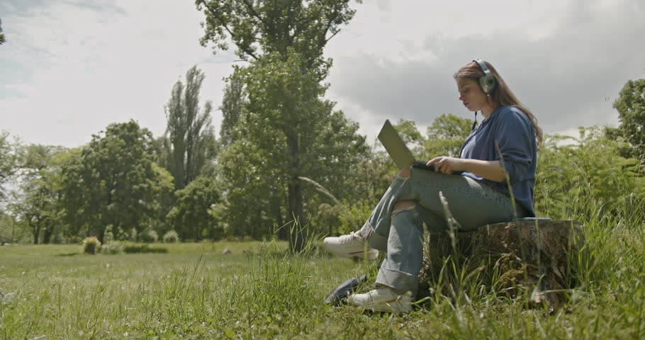Young woman working remotely from a park, enjoying the fresh air and nature sounds while using her laptop.