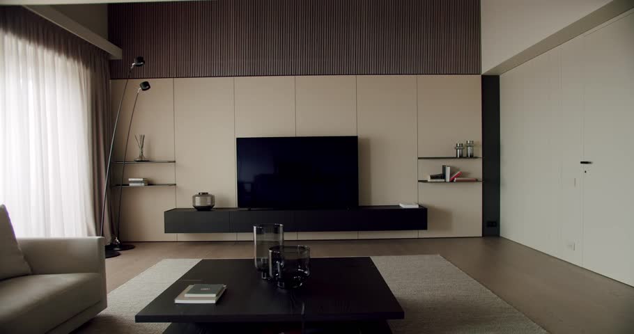 A stylish modern living room featuring a comfortable sofa, a wooden media unit, and a large flat-screen TV surrounded by tastefully minimalist decor and big bright windows.