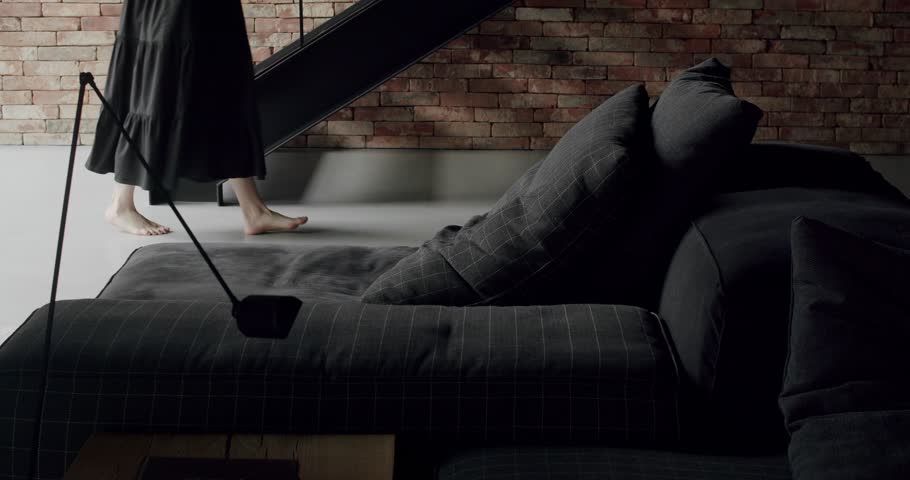 A woman in a long black dress was walking around the room. Modern contemporary Minimalist living room with bricks wall, large sliding windows and black furniture. Luxury Elegant Room