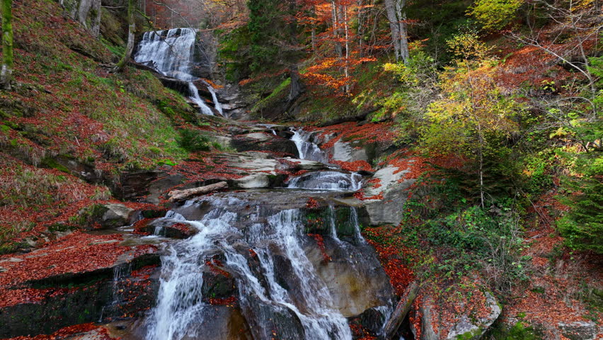 Golden autumn forest with a serene waterfall, silky water flowing over stones and vibrant leaves scattered across the peaceful scene. Aerial nature video.