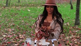 Beautiful young latin woman in a brown hat and jacket is throwing autumn leaves in the air in slow motion. She is sitting at a wooden table with a cup of coffee and an alarm clock - Powered by Shutterstock - Get 15% off with code: PIKWIZARD15