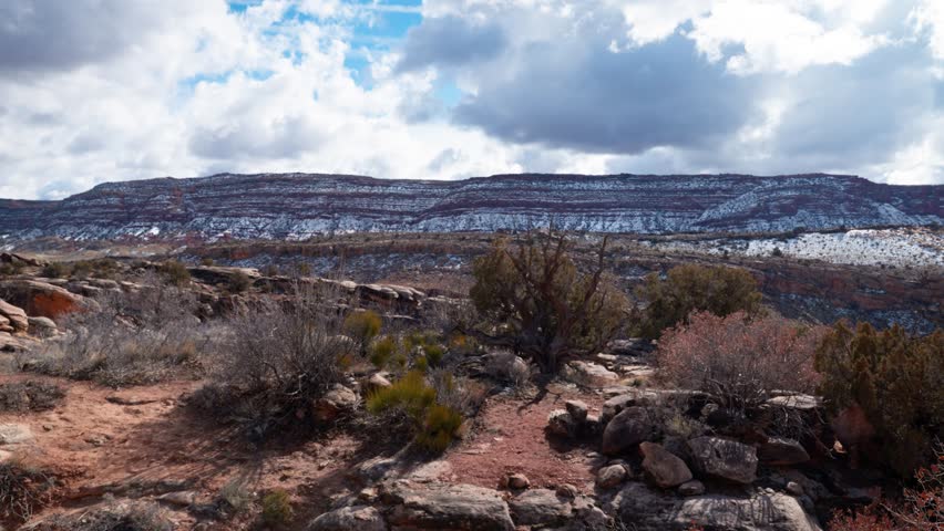 Static wide shot of large mountains covered in snow on a sunny day surrounded by shrubs in Arches National Park, Utah.
