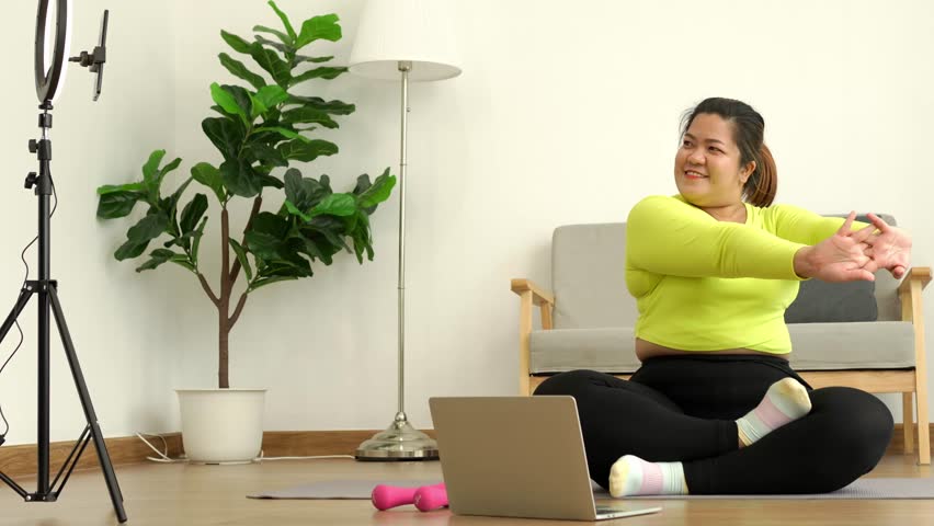 Asian overweight woman doing stretching exercise at home on fitness mat. Home activity training, online fitness class. Stretching training workout on yoga mat at home for good health and body shape.