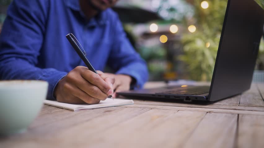 Close-up of a focused student or freelancer writing notes while working on a laptop outdoors. Unrecognizable individual in a casual setting taking notes by hand in a peaceful workspace.