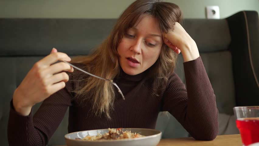 Young woman sits alone at a table, slowly eating her meal. Boredom and unhappiness are evident on her face as she engages in a solitary dining experience