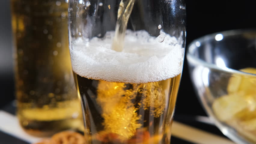 Pouring a cold beer from a bottle into a glass, creating a frothy head and bubbles rising. Nearby, a bowl filled with crispy potato chips and salty pretzels