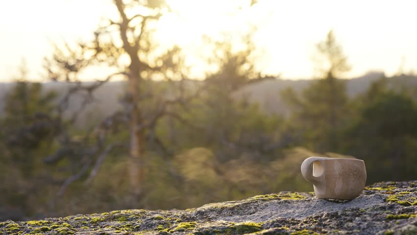 A traditional Finnish wooden kuksa cup on a gray rock in the forest, illuminated by the rays of the setting sun, with steam rising from the hot drink, coffee, tee inside