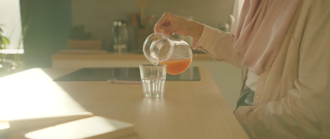 View on hands woman pouring orange juice from glass jar into glass at sunlight kitchen home. Camera moving around. 360 degrees