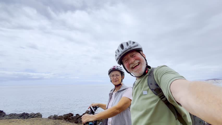 Two happy old mature people enjoying and riding bikes together to be fit and healthy outdoors. Active seniors having fun taking a selfie picture with camera