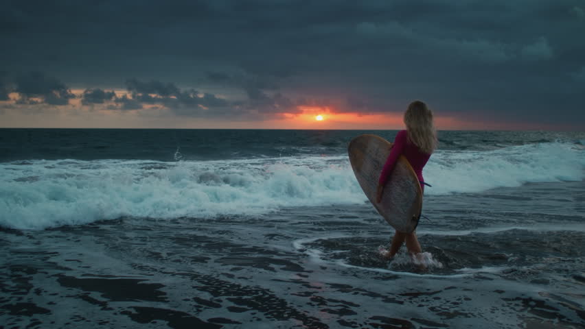Young woman surfer walks along sand ocean beach, holds surfboard above head after sup training. Attractive girl walking or run to surfing with board. Evening dusk on bali, indonesia. Pink swuimsuit