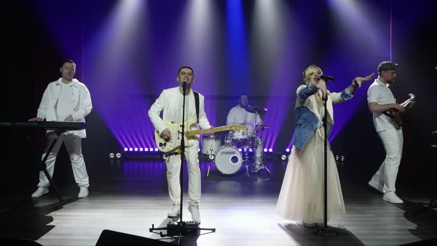 Cover band, dressed in white, performs on a stage lit with purple and yellow stage lights. The band consists of a drummer, guitarist, keyboardist, and a female singer