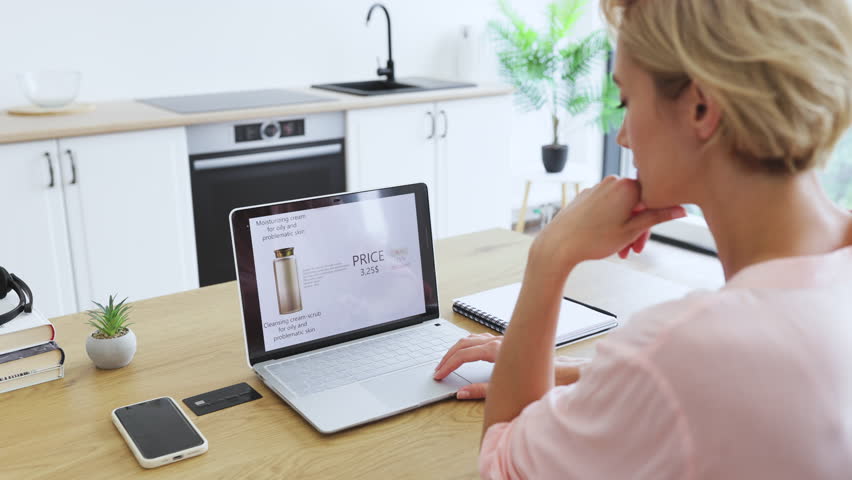 Caucasian woman using laptop to order cosmetics online in modern kitchen. Focused on delivery website compares prices and discounts. Casual and relaxed setting with smartphone and credit card nearby.