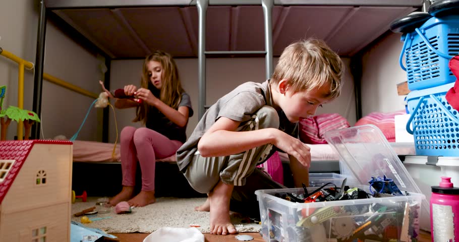Real time of concentrated girl looking down while sitting on bunk bed adjusting dress on small doll with hands in daylight and little brother sitting near on floor playing with toys in box small room