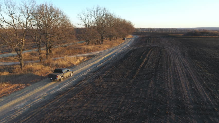 Aerial shot of car moving along road near plowed meadow at autumn. Flying over pickup truck driving through rural path near ploughed field. Off road vehicle moving on route near farmland at sunset