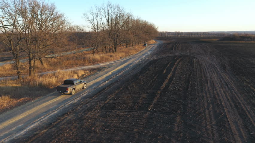 Aerial shot of car moving along road near plowed meadow at autumn. Flying over pickup truck driving through rural path near ploughed field. Off road vehicle moving on route near farmland at sunset
