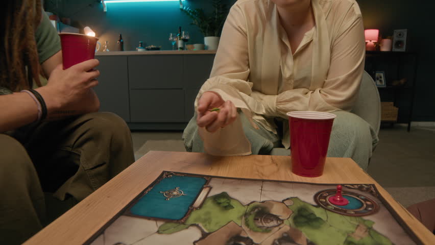 Zoom-out cropped slowmo shot of player rolling dice on map while playing board game together with friends in cozy home setting late in evening