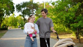 A happy brunette man in a gray T-shirt together with his wife who is holding their little daughter in a white dress are walking in the park together with a stroller for a child in the summer - Powered by Shutterstock - Get 15% off with code: PIKWIZARD15