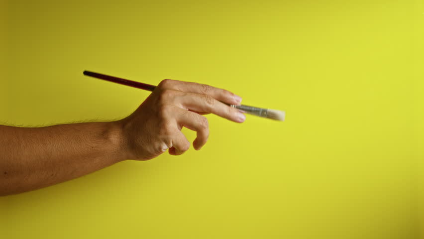 Hand holding paintbrush against a bright yellow background showcasing fingers in detail and painting concept