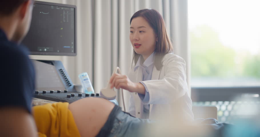 Asian Obstetrician Performs Ultrasound on Pregnant Patient, Explaining Baby Development. Modern Medical Equipment Displays Detailed Sonogram Images as Doctor Gestures Towards Screen