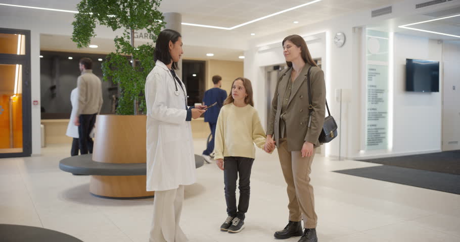 Active Modern Hospital Lobby with a Female Doctor Greeting Visitors, Providing Medical Information on a Tablet Computer. Healthcare Personnel Assisting Patient in Wheelchairs in the Background