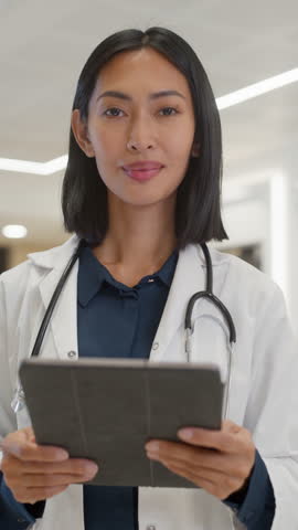 Vertical Screen: Young Asian Female Doctor in White Coat, with a Stethoscope Around Her Neck, Standing in a Hospital Hall and Using Tablet Computer. Medical Worker Looks at Camera with a Smile