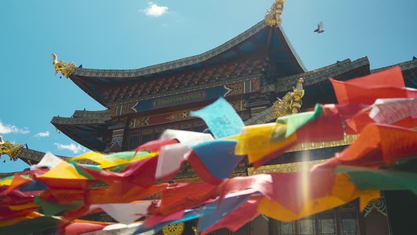Colorful prayer flags flutter in the foreground of an intricately designed Tibetan temple under a vibrant blue sky in Shangri-La City, blending culture and spirituality.