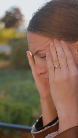 Young woman gently massaging temples with her fingers, her eyes closed as if she is trying to relieve headache or stress, she is outdoors in park. Close-up vertical video