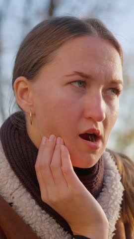 Young woman expressing various signs of pain and discomfort due to toothache. Facial expressions reveal intensity of suffering impact on her well-being. In park on bench in open air. Vertical video.