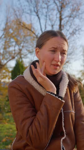 Young woman expressing various signs of pain and discomfort due to toothache. Facial expressions reveal intensity of suffering impact on her well-being. In park on bench in open air. Vertical video.