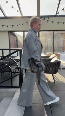 Stylish mature woman with short hair in grey suit with jacket and pants, accessorized and handbag walking indoors. Trendy outfit for successful women over 50 years.