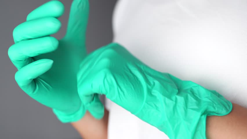 Young female doctor putting on green medical gloves before examining the patient on a grey background. Close up. Medicine, plastic surgery, health care concept