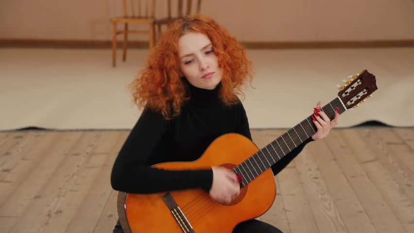Thoughtful attractive red-haired curly girl plays guitar sitting on stage in room. Girl guitarist creates music on guitar.