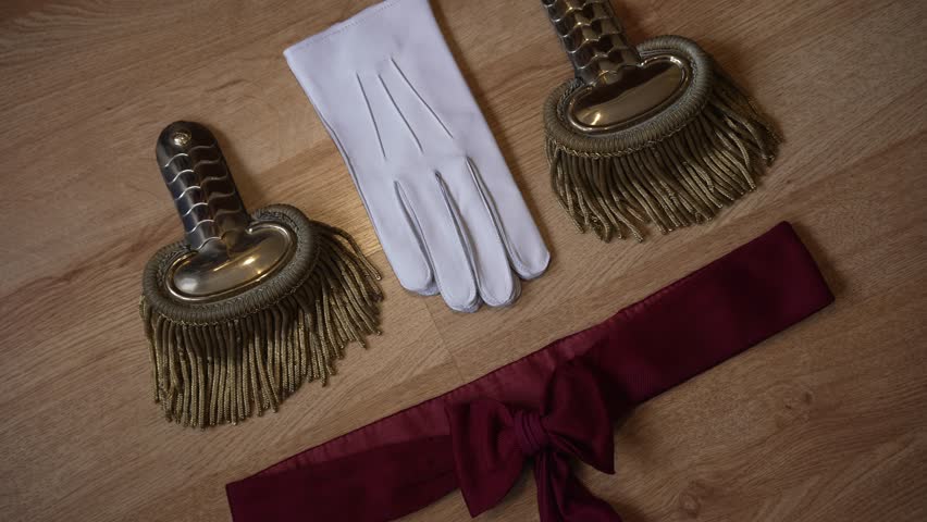 Elegant military accessories: gold epaulettes, white gloves, and a red sash, laid on wood
