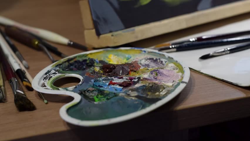 A female artist squeezes white acrylic paint onto her palette. Close-up shot. On the table beside her, brushes, palette knives, and other art tools are arranged.