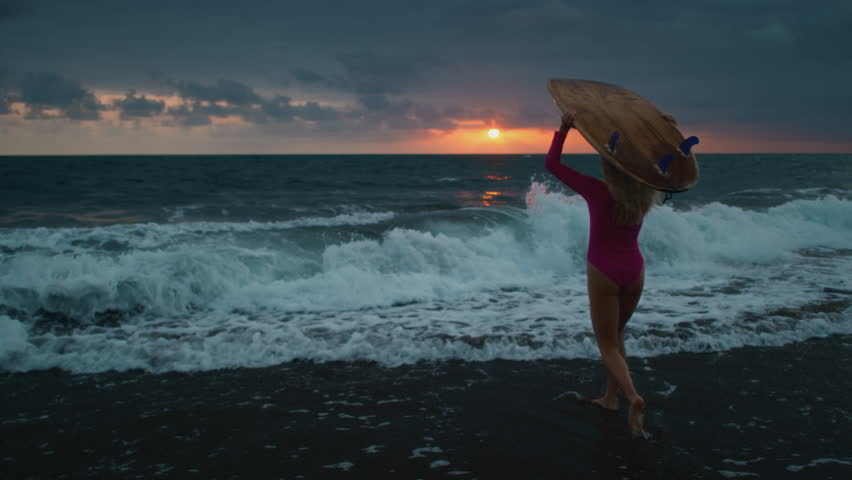 Young woman surfer walks along sand ocean beach, holds surfboard above head after sup training. Attractive girl walking or run to surfing with board. Evening dusk on bali, indonesia. Pink swuimsuit