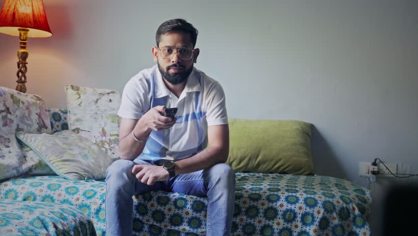Front view of an Indian adult man sitting on the sofa, using remote and watching TV.
