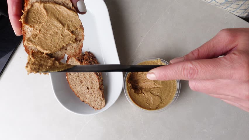 Hands spreading homemade chicken liver pate onto whole-grain bread, topped with fresh tomato slices and arugula. A rustic, savory snack with a touch of elegance and freshness.