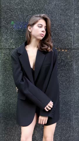 Stylish woman wearing elegant outfit oversize black blazer, beautiful model stands, poses on dark gray background. Modern high fashion, contemporary street style on minimalist backdrop