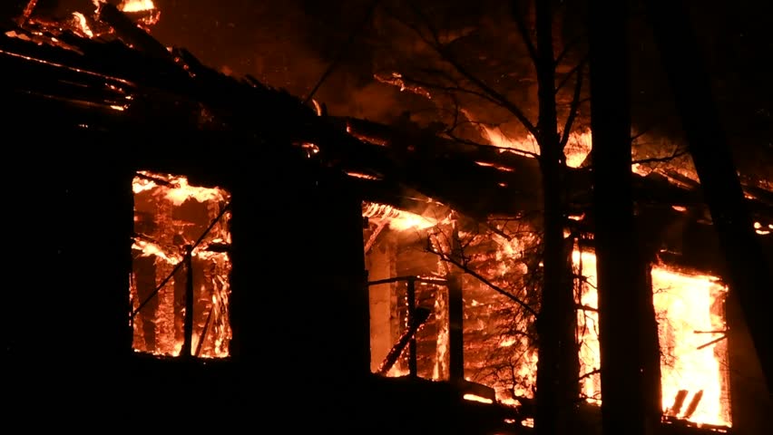 House is on fire at night, flames inside at home, burning windows. The wooden building is ablaze, completely engulfed in fire. Loss of housing, disaster, trouble for unhappy people.