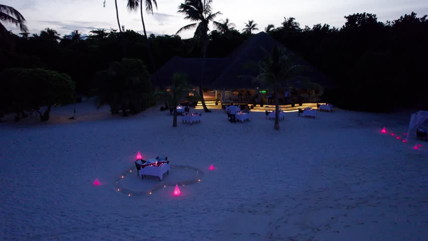 Romantic dinner by the ocean, white sand. Maldives. Lights and candles are burning. There are tables. Supper. Sunset with big clouds over the island. Pink backlight. Green palm trees. Turquoise water