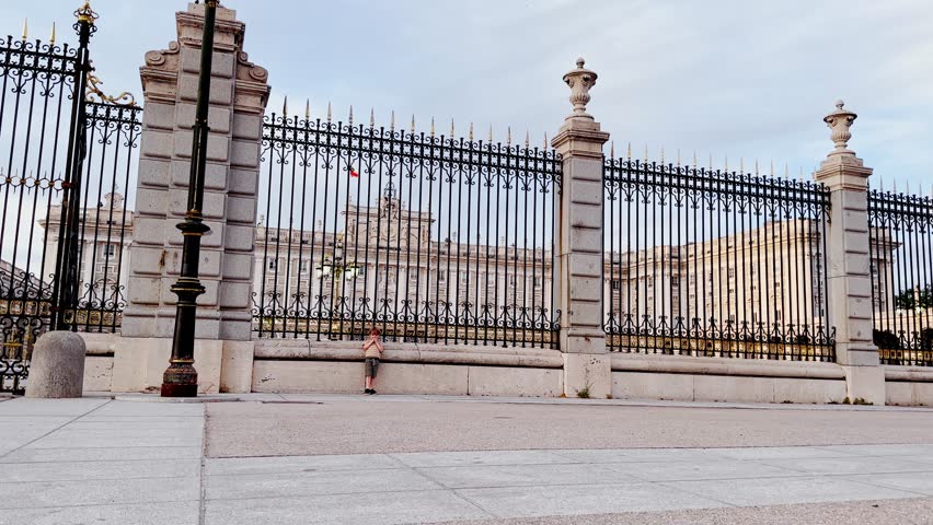 A young child in a striped shirt joyfully runs along a spacious courtyard of Royal palace of Madrid with an ornate iron fence behind him