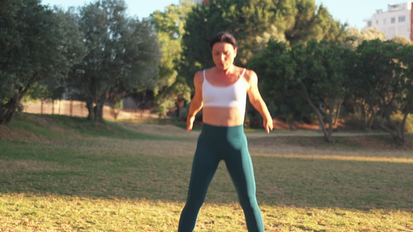 Sporty woman practicing jumping squats in city park on sunny day. Young fit woman in sportswear exercising on lawn during daytime