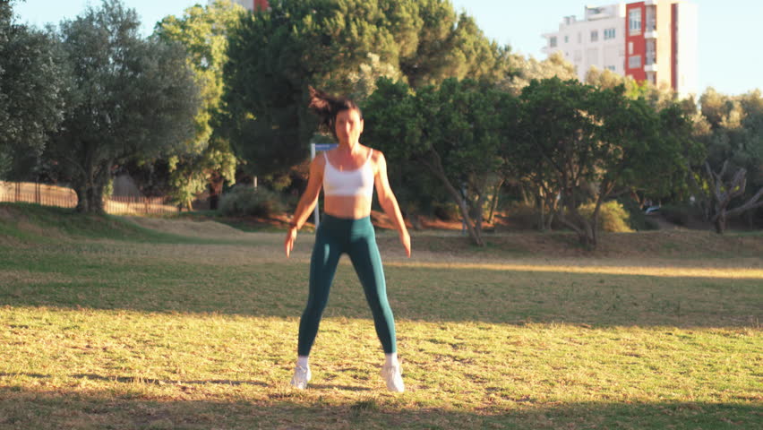 Sporty woman practicing jumping squats in city park on sunny day. Young fit woman in sportswear exercising on lawn during daytime